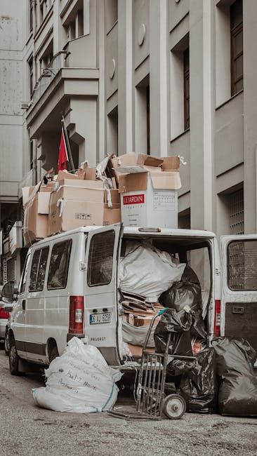A narrow urban alleyway cluttered with a large wheeled rubbish cart positioned in the foreground, made of sturdy fabric with visible creases and discolouration, indicating frequent use. Behind the car