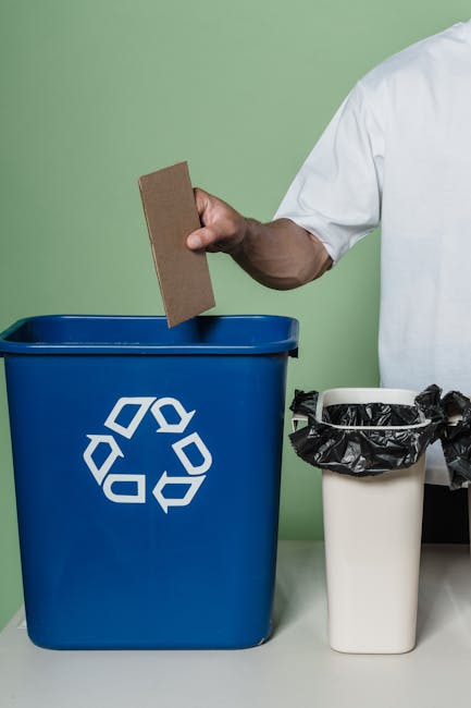 Two clear plastic recycling bins with green recycling symbols on the front, placed on a light grey concrete surface, with one bin labeled 'GLASS' in white text on a red background. The bins are intend